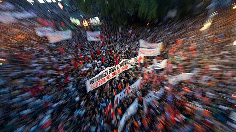 Multitudinaria manifestación en la calle con efecto de desenfoque radial, donde un grupo porta una pancarta blanca con texto en catalán y banderas.