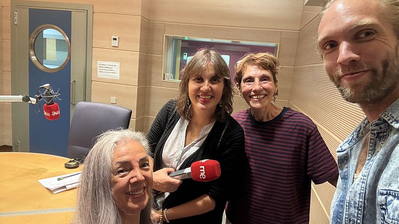Selfi de cuatro personas sonrientes en un estudio de radio, con la periodista Olga Baeza en el centro sosteniendo un micrófono de RNE y rodeada de paneles acústicos.
