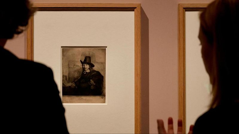 Interior de una galería de arte donde dos personas observan un grabado monocromático iluminado en la pared.