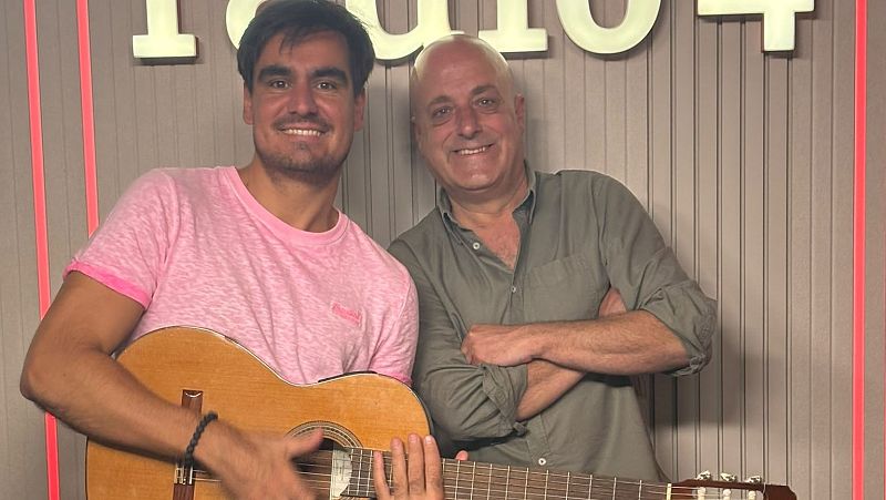Dos hombres sonrientes en un estudio de radio, uno sostiene una guitarra cl�sica y el otro posa con los brazos cruzados ante un panel con luz roja.