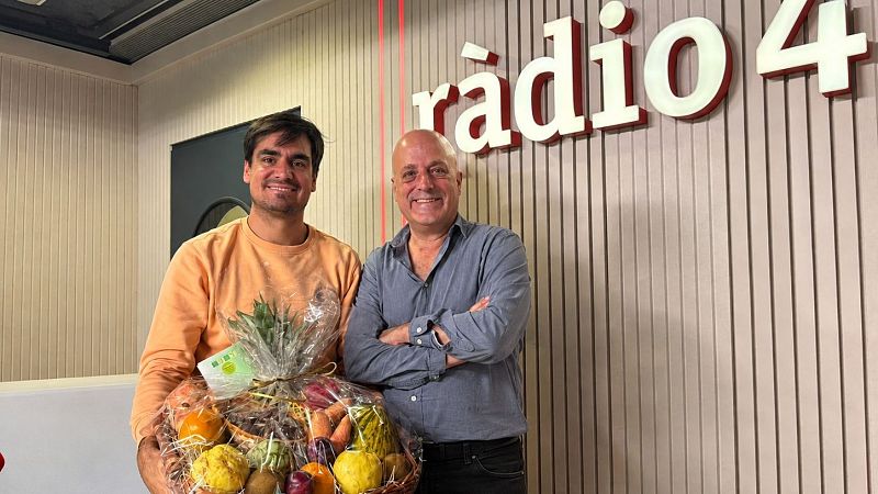 Dos hombres sonrientes en un estudio de radio; uno sostiene una cesta de mimbre llena de frutas y verduras, y el otro posa con los brazos cruzados.
