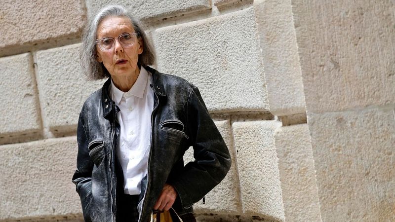 Plano medio de la escritora Anne Carson con cabello canoso y gafas, vistiendo camisa blanca y cazadora de cuero ante una pared de piedra.