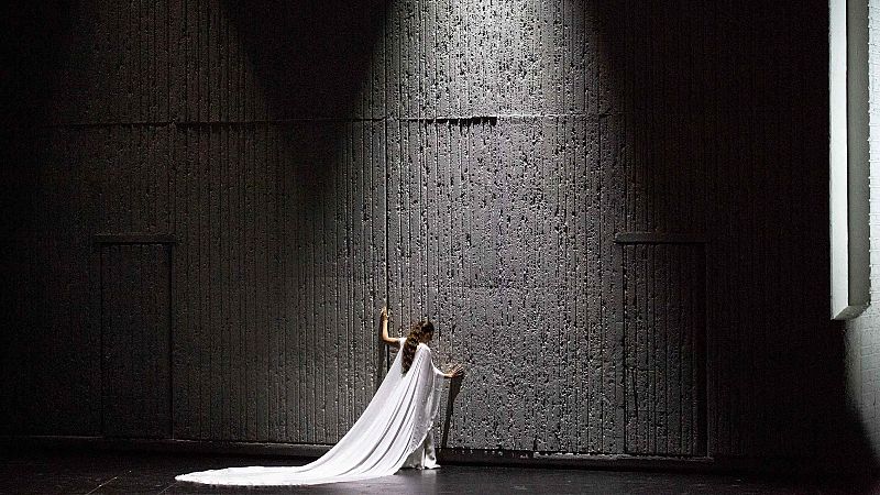 Mujer vestida de blanco con una larga capa apoyada contra un muro de hormig�n brutalista bajo una luz cenital en el escenario.