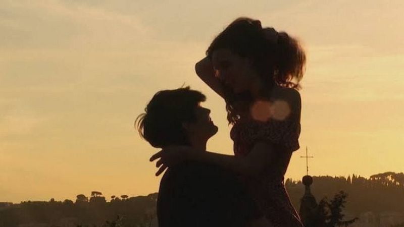 Fotograf�a a contraluz al atardecer de una pareja joven en silueta, donde un chico sostiene en alto a la chica frente a un cielo dorado y la silueta de una c�pula.