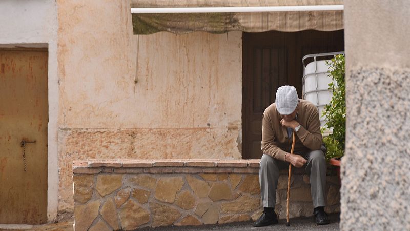 Un anciano, con gorra y camisa marrón, se sienta en un muro de piedra frente a una casa con puerta de madera y ventana con persiana. La imagen transmite una sensación de tristeza y soledad en un entorno rural.