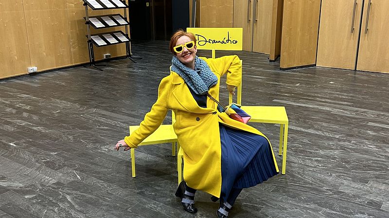La figurinista Guadalupe Valero posa sentada en sillas amarillas en un vest�bulo, vistiendo un abrigo amarillo, falda azul y gafas de sol, sonriendo ante la c�mara.