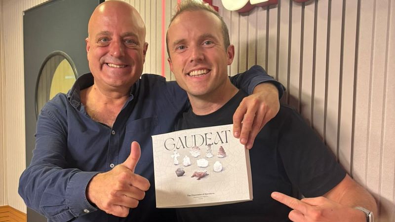 Dos hombres sonrientes en un estudio de radio, uno muestra una caja de bombones blanca con la palabra GAUDEAT y fotograf�as de dulces inspirados en Gaud�.