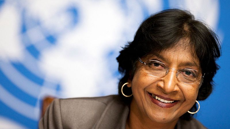 Retrato de la jurista Navi Pillay sonriendo con chaqueta formal, gafas y el emblema de la ONU de fondo.