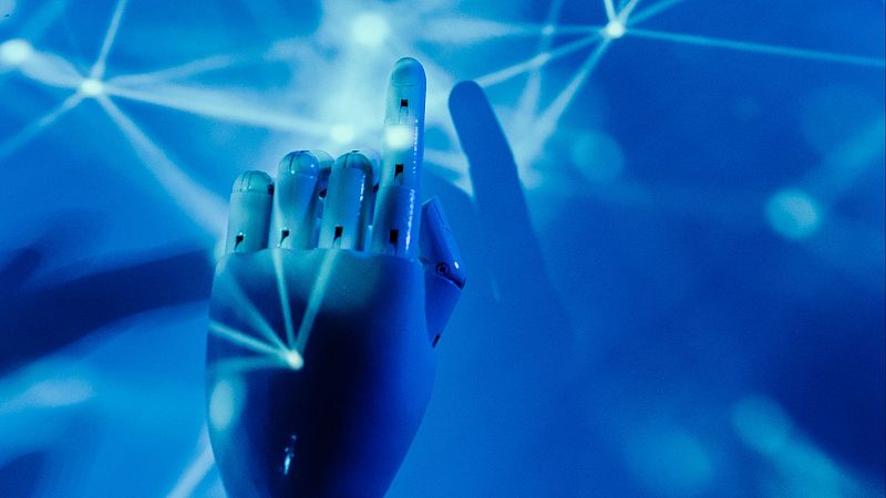 Mano robótica de diseño tecnológico y color claro con el dedo índice apuntando hacia arriba sobre un fondo azul con líneas luminosas.