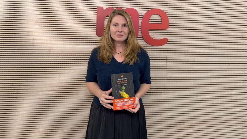 La escritora Tatiana Țîbuleac sostiene su libro con faja naranja frente a un panel de madera y el logotipo de RNE.