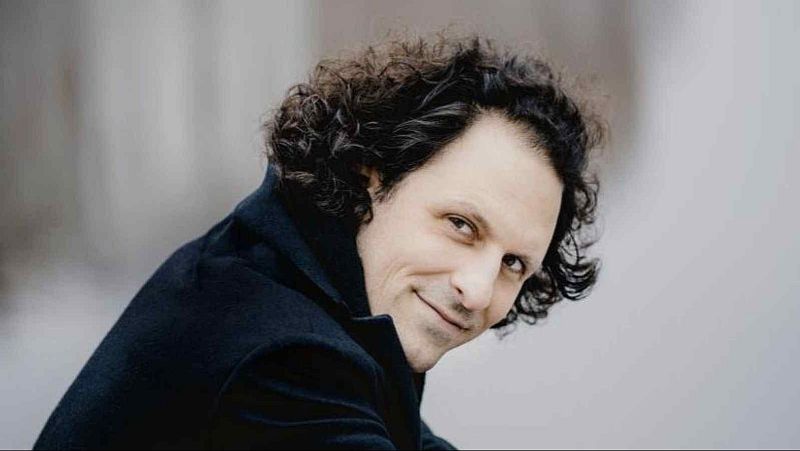 Primer plano del director de orquesta Omer Meir Wellber mirando hacia atr�s con una sutil sonrisa, vistiendo una chaqueta oscura sobre fondo difuminado.