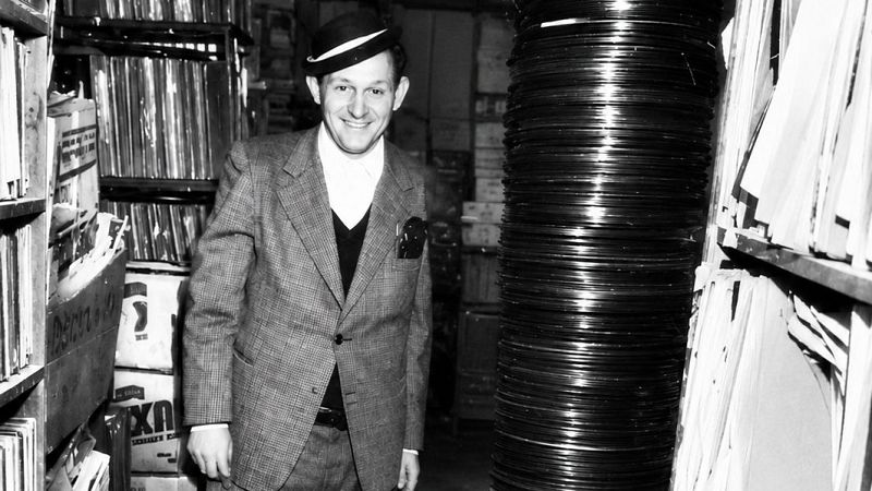 Hombre sonriente con traje a cuadros y sombrero junto a una gran torre de discos de pizarra en un archivo musical.