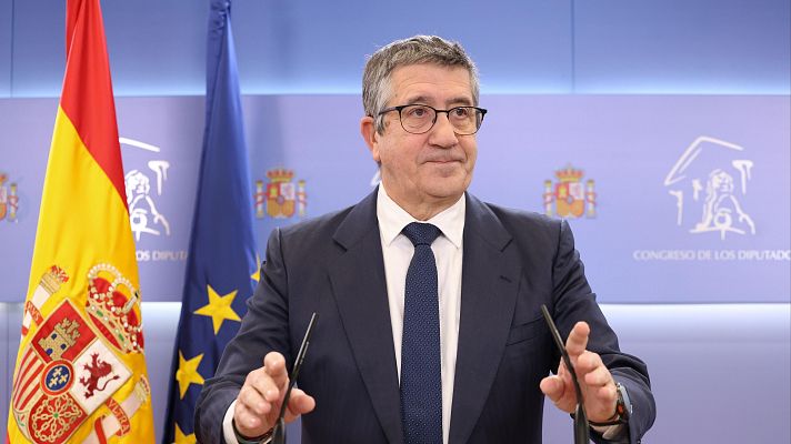 Primer plano de Patxi López en una sala institucional, con las banderas de España y la UE al fondo. Lleva traje oscuro, camisa blanca y corbata azul, y se apoya en un atril.
