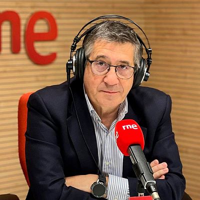 Primer plano de Patxi López en una sala institucional, con las banderas de España y la UE al fondo. Lleva traje oscuro, camisa blanca y corbata azul, y se apoya en un atril.