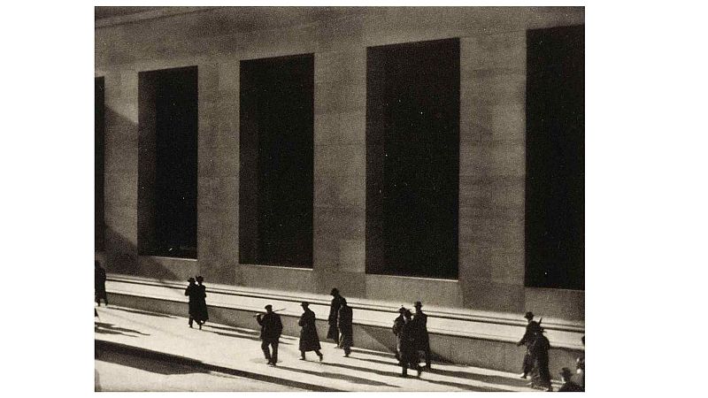 Fotograf�a en blanco y negro de 1915 que muestra la fachada monumental de un edificio con grandes ventanales y transe�ntes con abrigos y sombreros proyectando largas sombras.