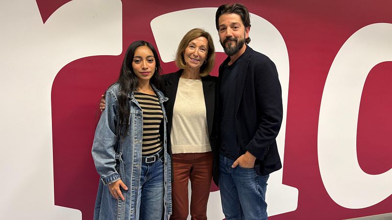 Tres personas posan sonrientes ante un mural de RNE, con la periodista Yolanda Flores en el centro abrazando al actor Diego Luna y a una joven.