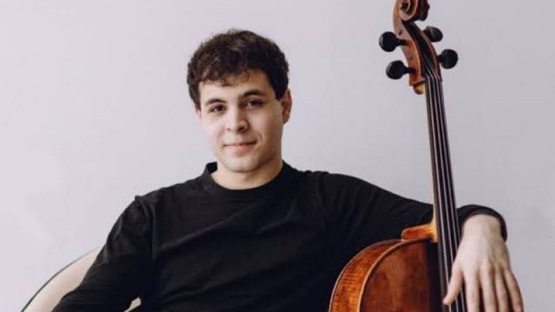 Retrato en plano medio del violonchelista Luis Aracama posando sonriente junto a su instrumento sobre fondo gris claro.