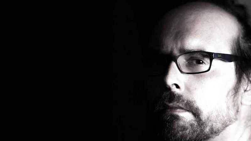 Primer plano de un hombre adulto con gafas y barba, iluminado con un intenso claroscuro sobre fondo negro.