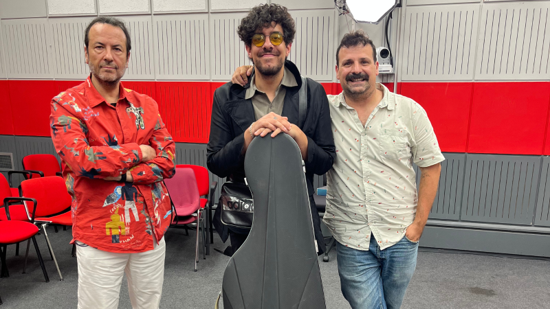 Tres hombres posan en un estudio de grabaci�n con paneles ac�sticos: Ram�n Gener con camisa roja, un joven con gafas amarillas apoyado en una funda de instrumento, y Carles Mesa con camisa clara.