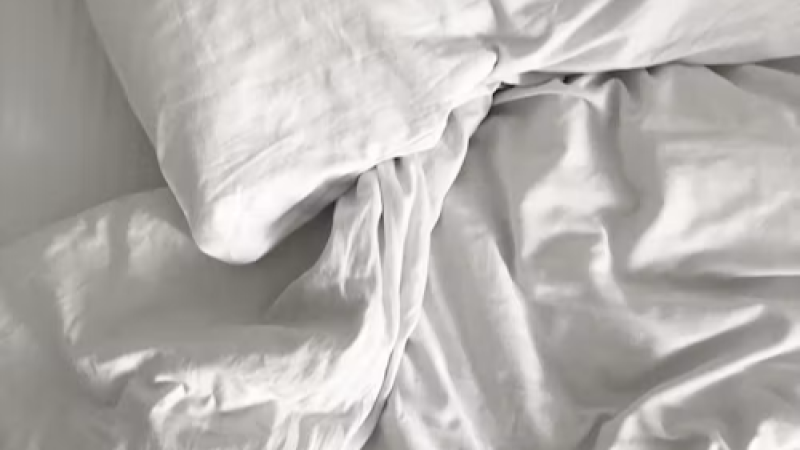 Plano cercano de una cama deshecha con ropa de cama blanca, mostrando una almohada arrugada y un edred�n lleno de pliegues bajo una luz suave.