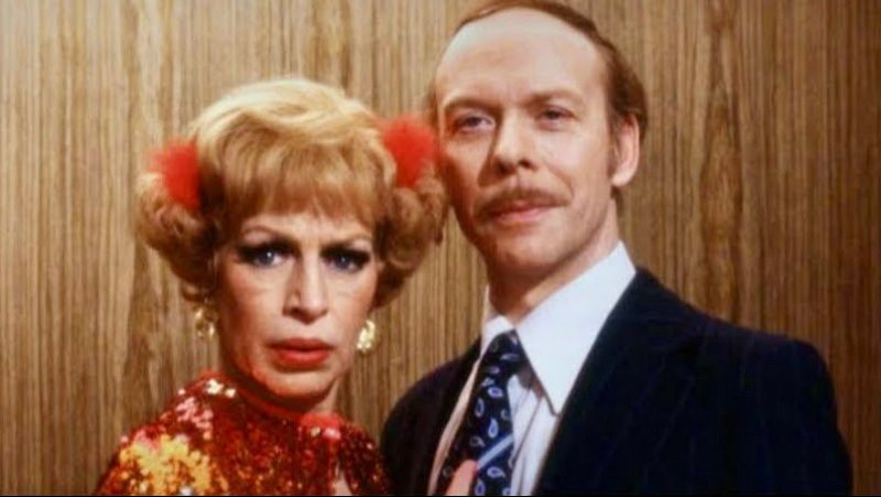 Plano medio de dos actores caracterizados de �poca, ella con vestido de lentejuelas y pompones rojos en el cabello, y �l con traje formal y bigote.
