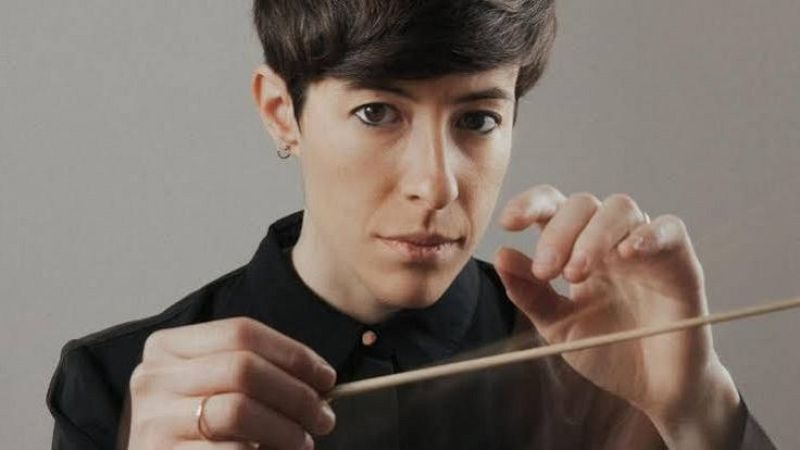 La directora de orquesta Isabel Rubio posa seria en primer plano, sosteniendo una batuta de madera con las manos a la altura del pecho.