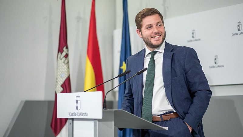 Nacho Hernando sonríe en una comparecencia institucional ante un atril con el escudo de Castilla-La Mancha y tres banderas oficiales al fondo.