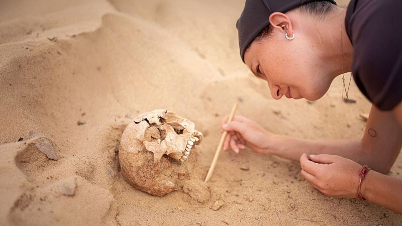 Una arqueóloga con gorra negra retira arena con una vara para desenterrar un cráneo humano fósil en un terreno desértico.