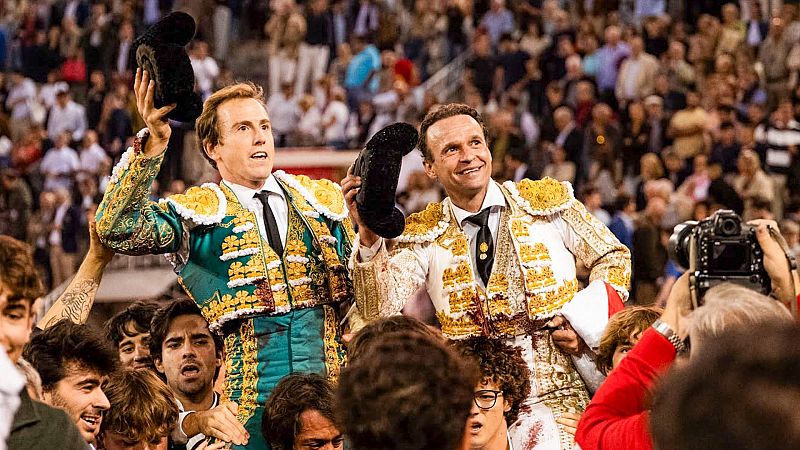 Dos toreros con trajes de luces verde y blanco son llevados a hombros por aficionados en una plaza de toros ante una multitud entregada.