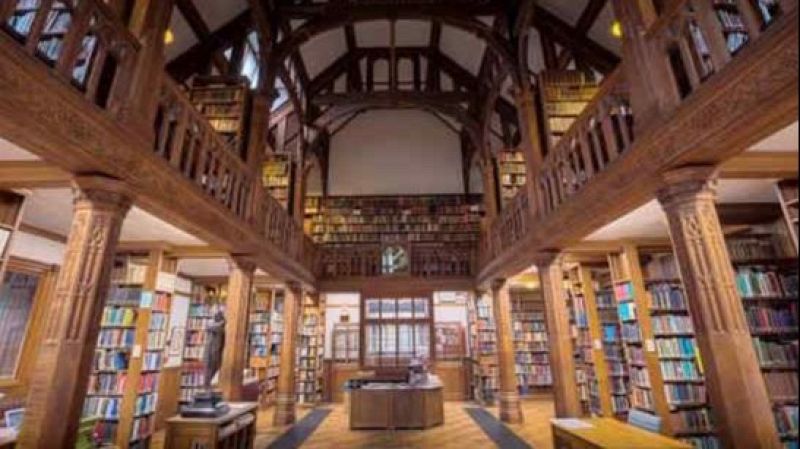 Interior de una biblioteca hist�rica de dos plantas con estanter�as de madera noble, galer�a superior y un pasillo central iluminado con suavidad.