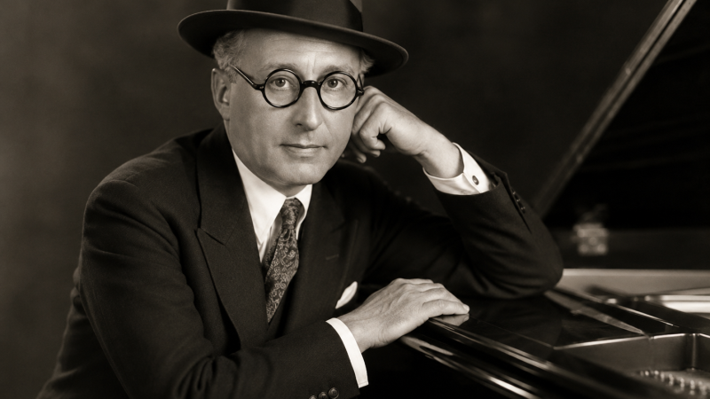 Retrato sepia de estilo cl�sico de los a�os 30 en el que un hombre con traje formal, sombrero y gafas redondas posa junto a un piano de cola brillante.