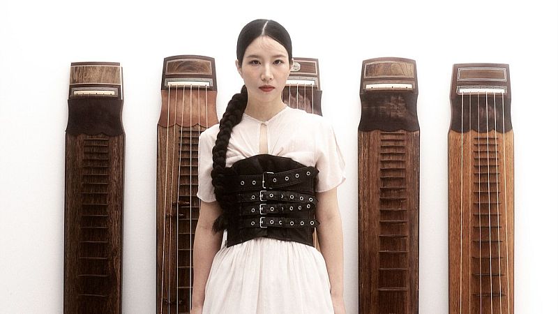 La artista surcoreana Hwang Gina posa seria ante un fondo blanco, vistiendo un cors� de cuero negro con hebillas sobre ropa clara y con el cabello en una larga trenza.