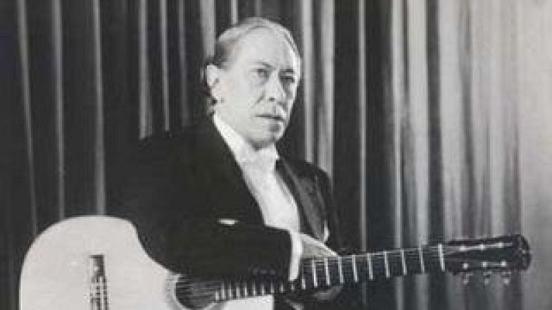 Fotograf�a en blanco y negro del guitarrista Agust�n Barrios con traje formal, posando seriamente en un estudio con su guitarra cl�sica en primer plano.