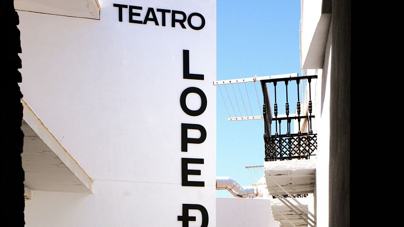 Calle estrecha y soleada entre edificios blancos con un r�tulo negro de �TEATRO� y un balc�n de forja bajo un cielo azul.