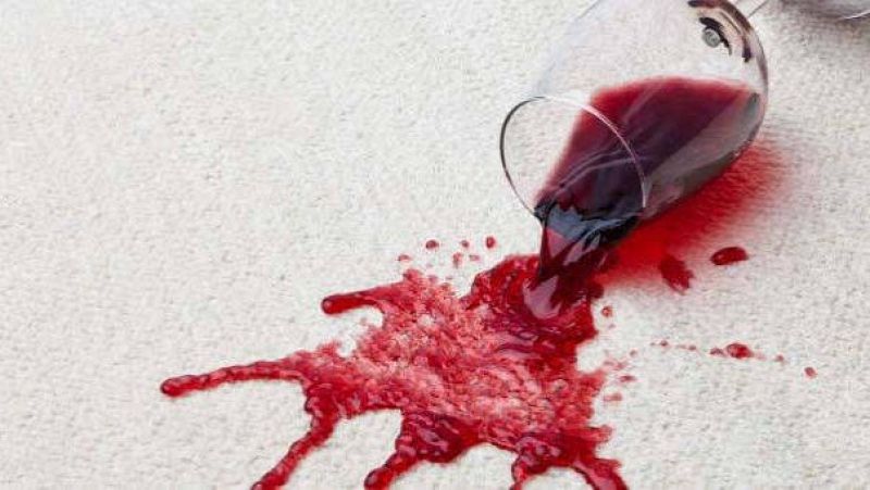 Copa de cristal tumbada sobre una alfombra clara de la que se derrama vino tinto formando una mancha.