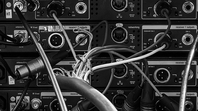 Primer plano en blanco y negro de la parte trasera de equipos de audio profesional con múltiples conexiones y cables.