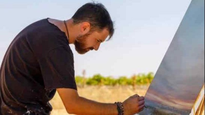 Joven pintor de perfil con barba y camiseta negra pinta un atardecer en un lienzo apoyado en un caballete al aire libre.
