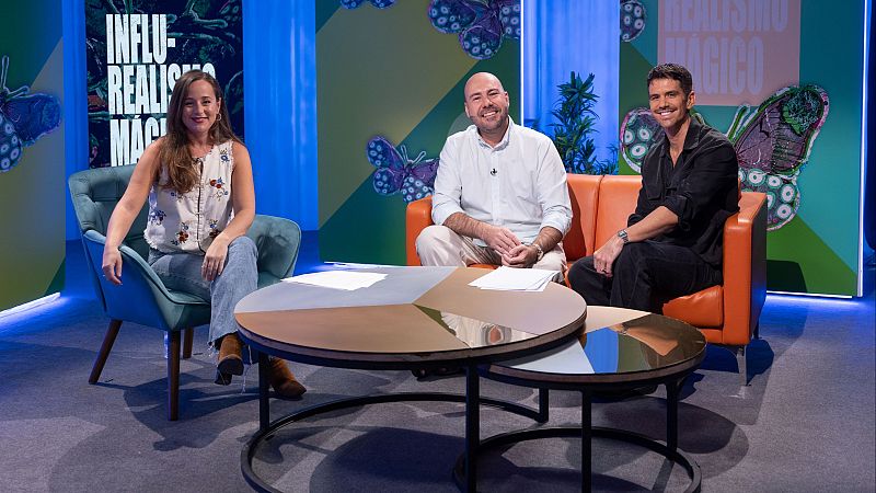 Tres personas sonrientes, una mujer y dos hombres, participan en una entrevista en un plat� decorado con paneles coloridos, mariposas y carteles con la leyenda Influ-Realismo M�gico.