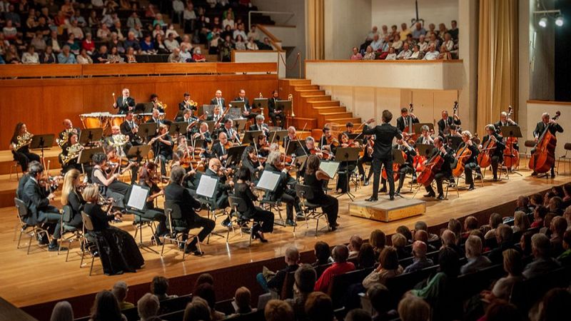 Concierto de m�sica cl�sica en un auditorio con m�sicos de negro dirigidos de espaldas y p�blico en la sala.