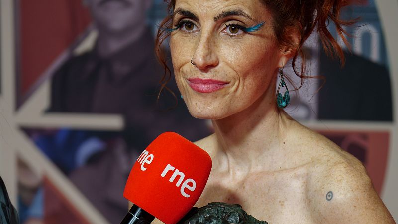 Primer plano de la actriz Miriam Garlo con cabello pelirrojo, maquillaje azul y sujetando una estatuilla de los Premios Goya junto a un micrófono de RNE.