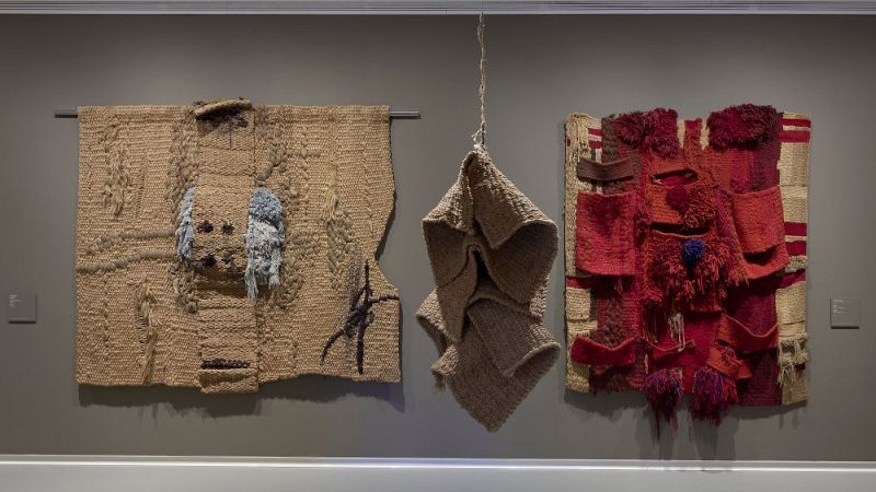 Sala de exposiciones con tres obras de arte textil contemporáneo sobre pared gris iluminadas por focos.
