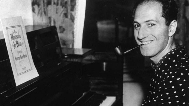 Fotograf�a en blanco y negro de George Gershwin tocando el piano, sonriendo con una pipa y con una partitura de Rhapsody in Blue en el atril.