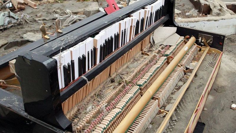 Piano vertical negro muy da�ado y cubierto de barro entre escombros, con el mecanismo interno expuesto y teclas rotas.