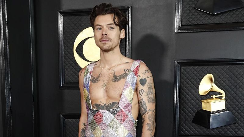 En la imagen, un hombre con tatuajes en pecho y brazos, posa con un mono estampado frente a un fondo oscuro. Se observan premios enmarcados.