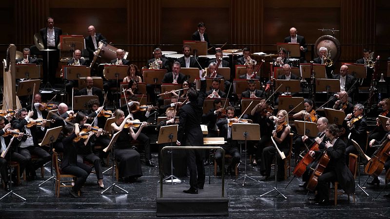 Orquesta sinfónica actuando en directo sobre un escenario de madera bajo una iluminación tenue, con el director de espaldas sosteniendo la batuta y músicos con instrumentos formales.