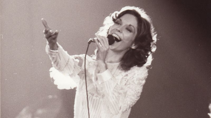 Karen Carpenter, en blanco y negro, canta al micrófono en un escenario con iluminación enfocada. Lleva una blusa blanca de manga larga con detalles. Expresión alegre.