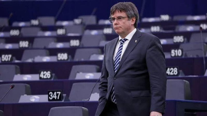 Carles Puigdemont camina serio por el hemiciclo del Parlamento Europeo con traje azul y un pin de lazo amarillo, rodeado de butacas vac�as.
