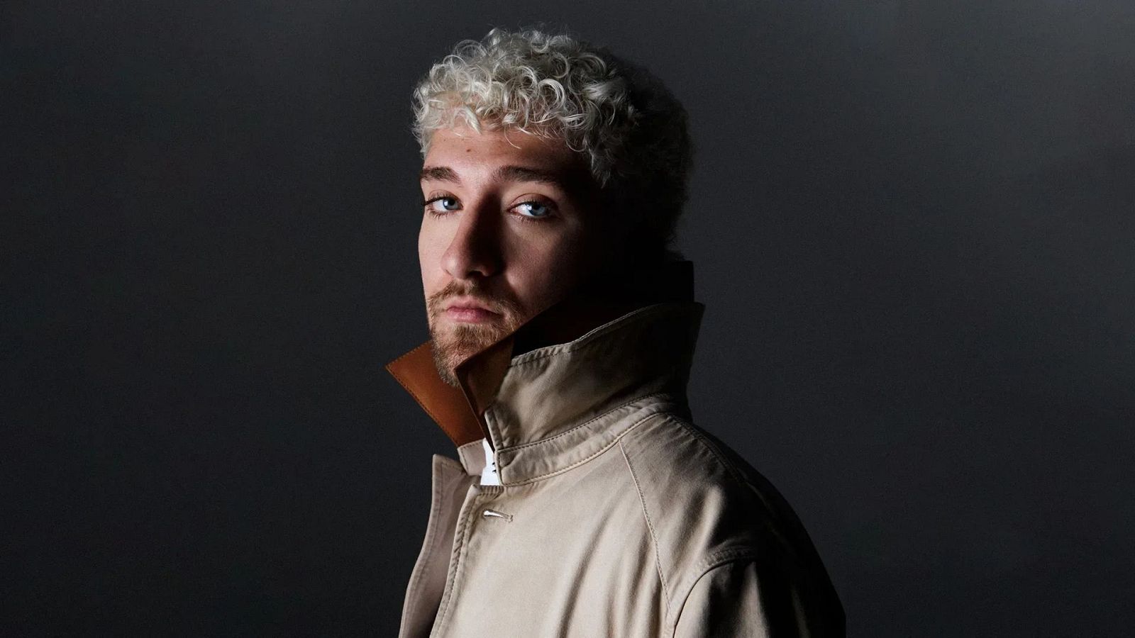 Plano medio corto de un hombre joven con cabello rizado rubio platino y barba corta, mirando fijamente a la c�mara con abrigo beige y una iluminaci�n de claroscuro sobre fondo oscuro.