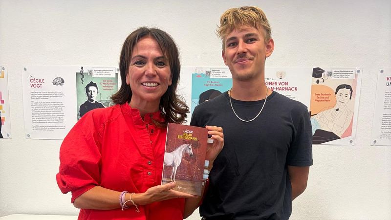 La periodista Susana Santaolalla y el escritor Nelio Biedermann posan juntos sosteniendo el libro Lázár ante una pared blanca con carteles.