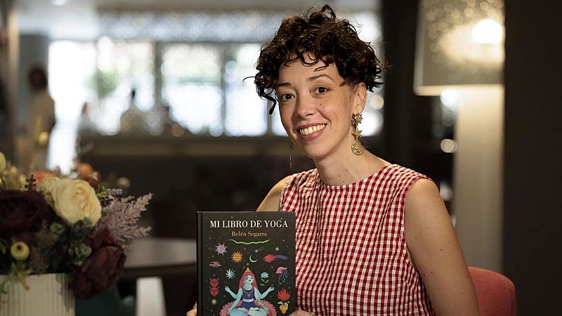 Plano medio de la ilustradora Belén Segarra sonriendo y sosteniendo su libro de yoga, vistiendo un top de cuadros vichy y pendientes dorados.